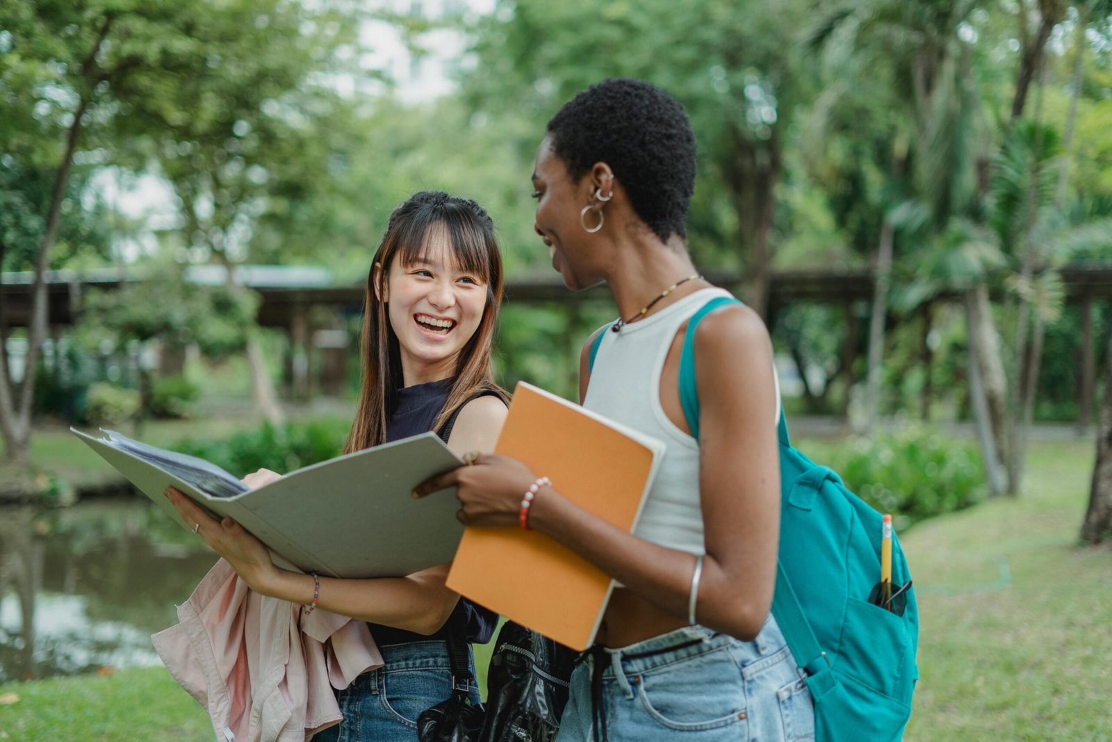 Young multiracial women in casual summer outfits with backpack speaking with each other while walking on grass with notepad and folder and looking at each other